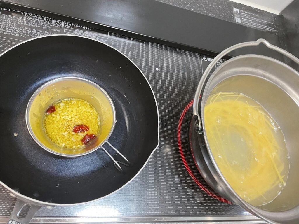 みじん切りにんにくと鷹の爪をオリーブオイルで弱火加熱中。IHコンロ対策でシェラカップを活用